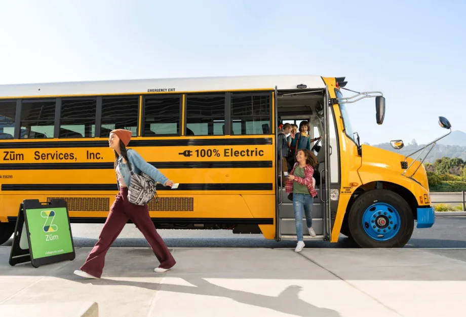 Students getting off a yellow electric school bus labeled “100% Electric” with Zūm Services branding