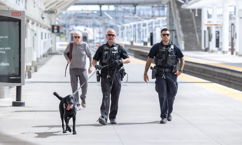 Two RTD transit police officers walk along a track with a police dog on a leash.