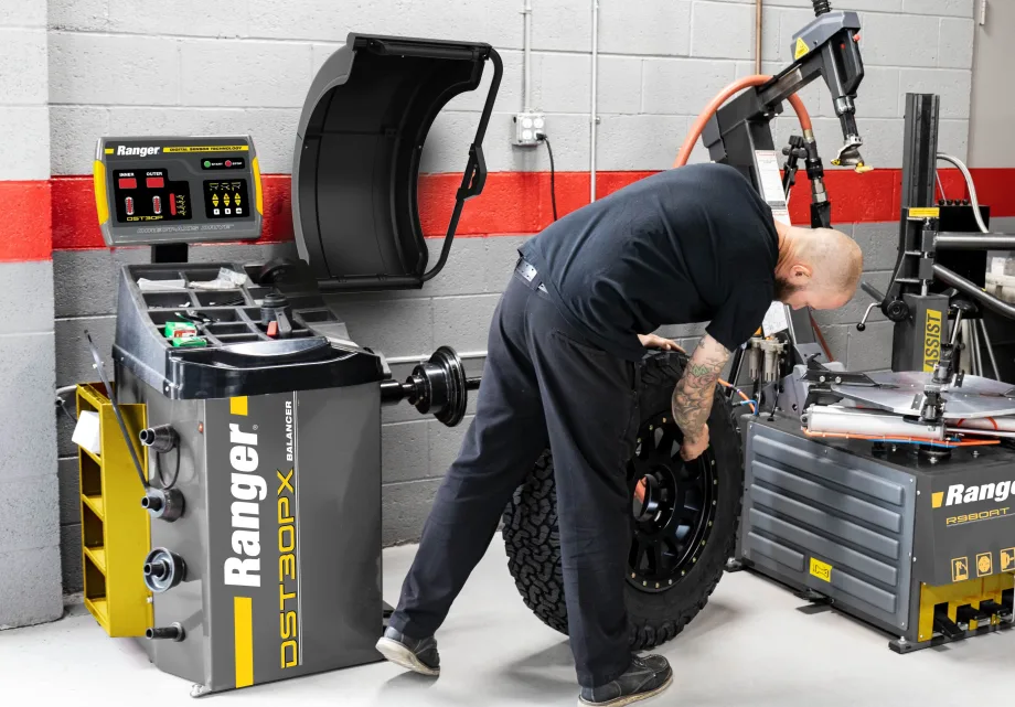 Automotive technician handling a large tire and wheel assembly beside a Ranger DST30PX wheel balancer and tire-changing equipment in a professional repair shop.